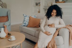 Smiling beautiful pregnant mixed race woman sitting on couch and touching her belly