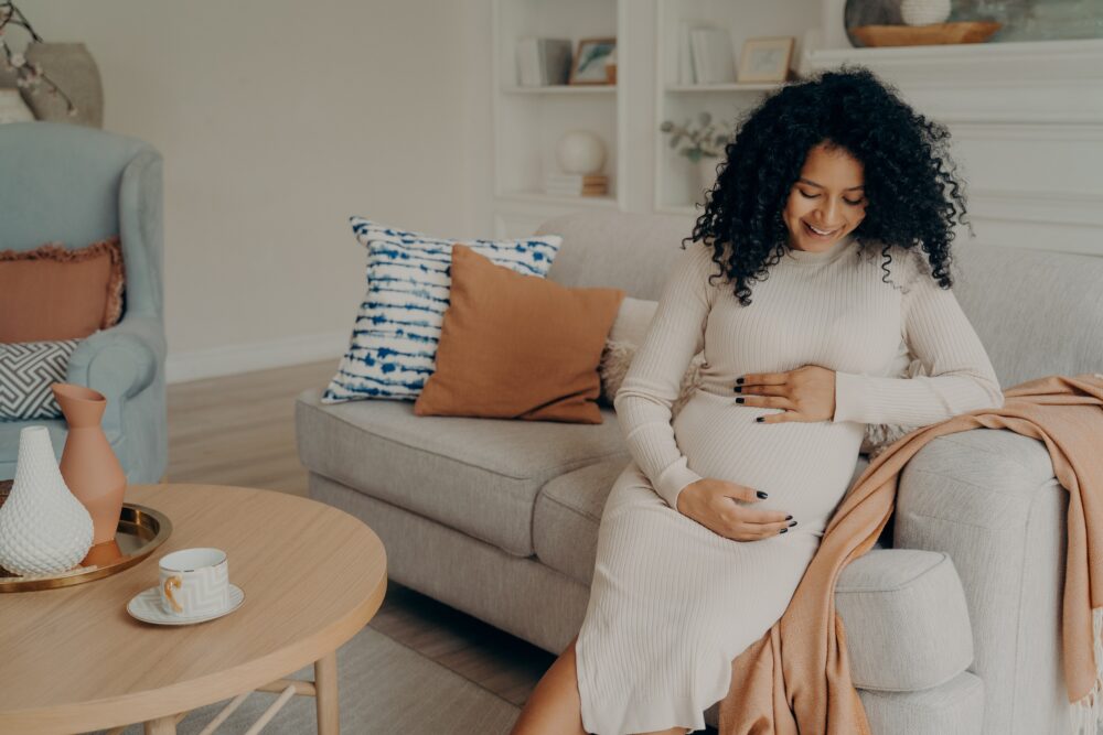 Smiling beautiful pregnant mixed race woman sitting on couch and touching her belly